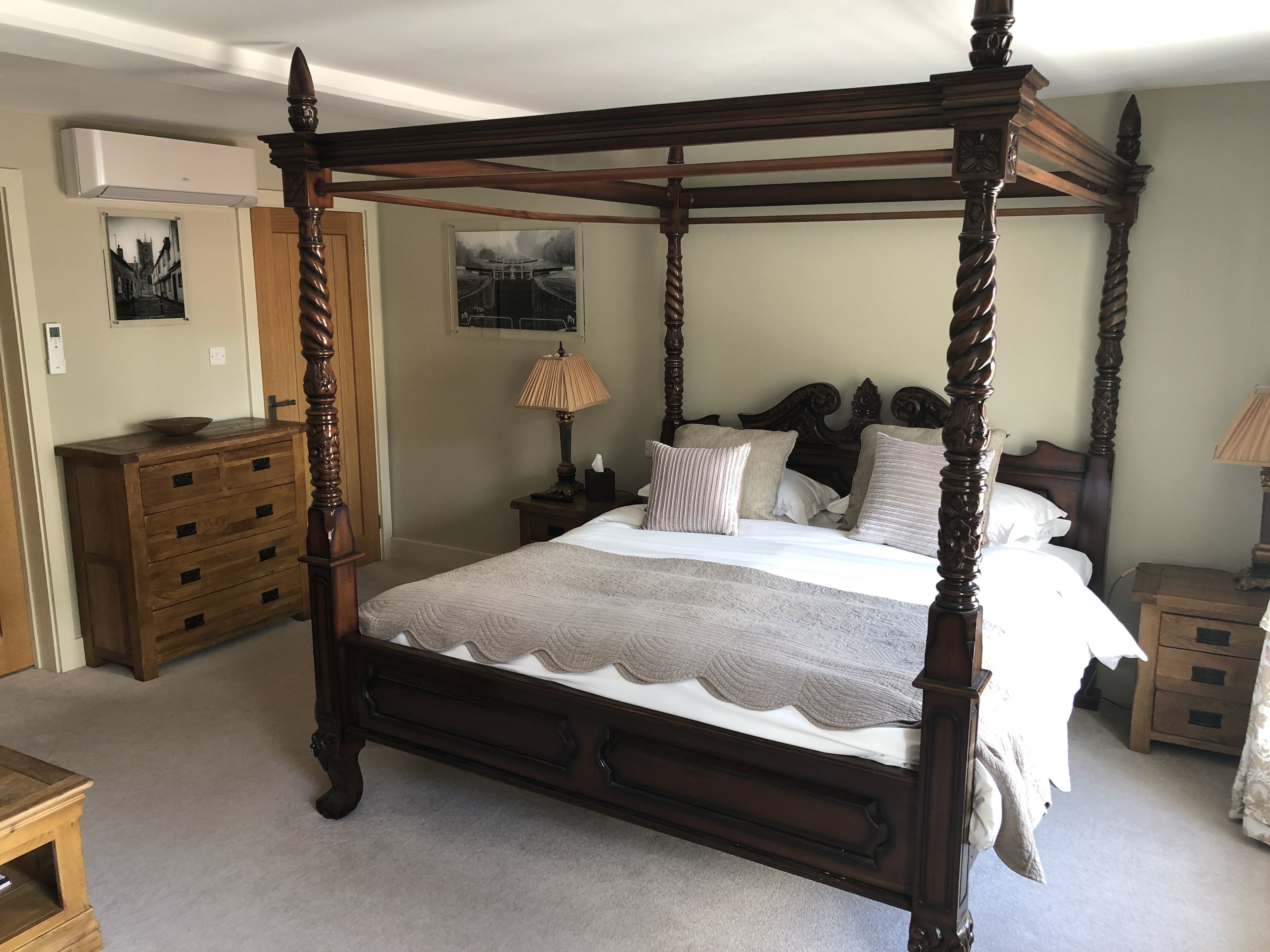 Un lit à baldaquin avec une courtepointe, une commode en bois, une table de chevet avec une lampe et un climatiseur fixé au mur.