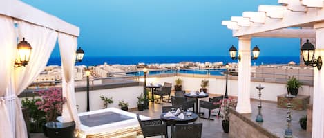 A rooftop terrace with tables and chairs, potted plants, and a view of the sea and cityscape.
