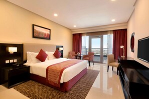 A hotel room with a large bed, a desk with a chair, a television, and a view of the outdoors. - Ramada Hotel & Suites by Wyndham Amwaj Islands Manama (Amwaj Islands)