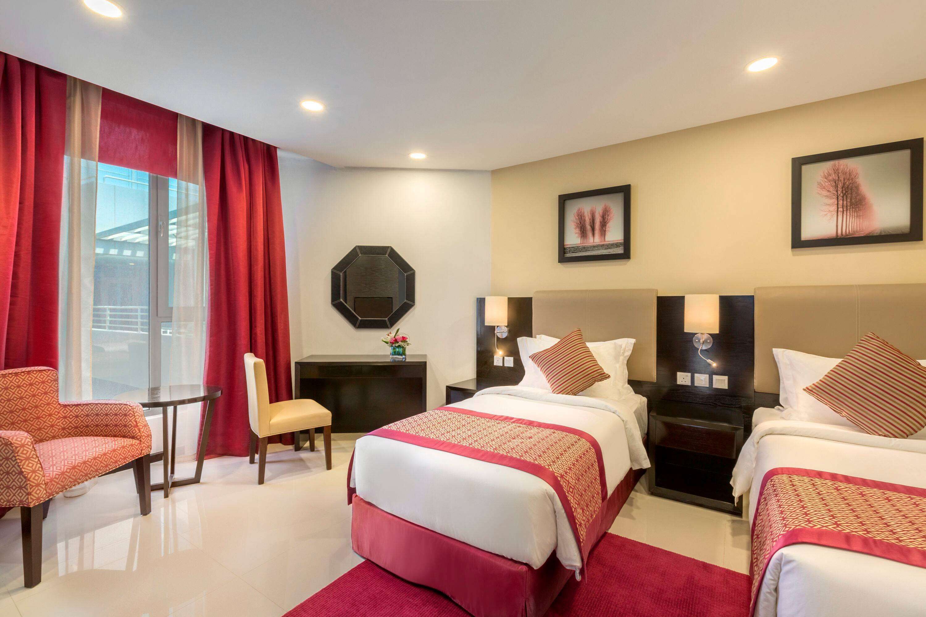 A hotel room with two beds, a desk, a chair, and a large window with red curtains.