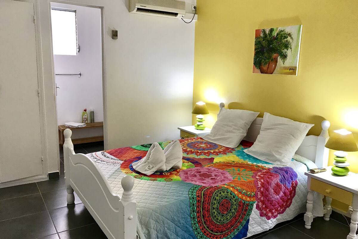 A bedroom with a bed, two bedside tables, a nightstand, a wall-mounted air conditioner, a door, and a wall painting.