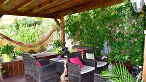 A covered patio with wicker furniture, a hammock, and lush greenery. - PARADIS TROPICAL appart'hotel (Baillif)