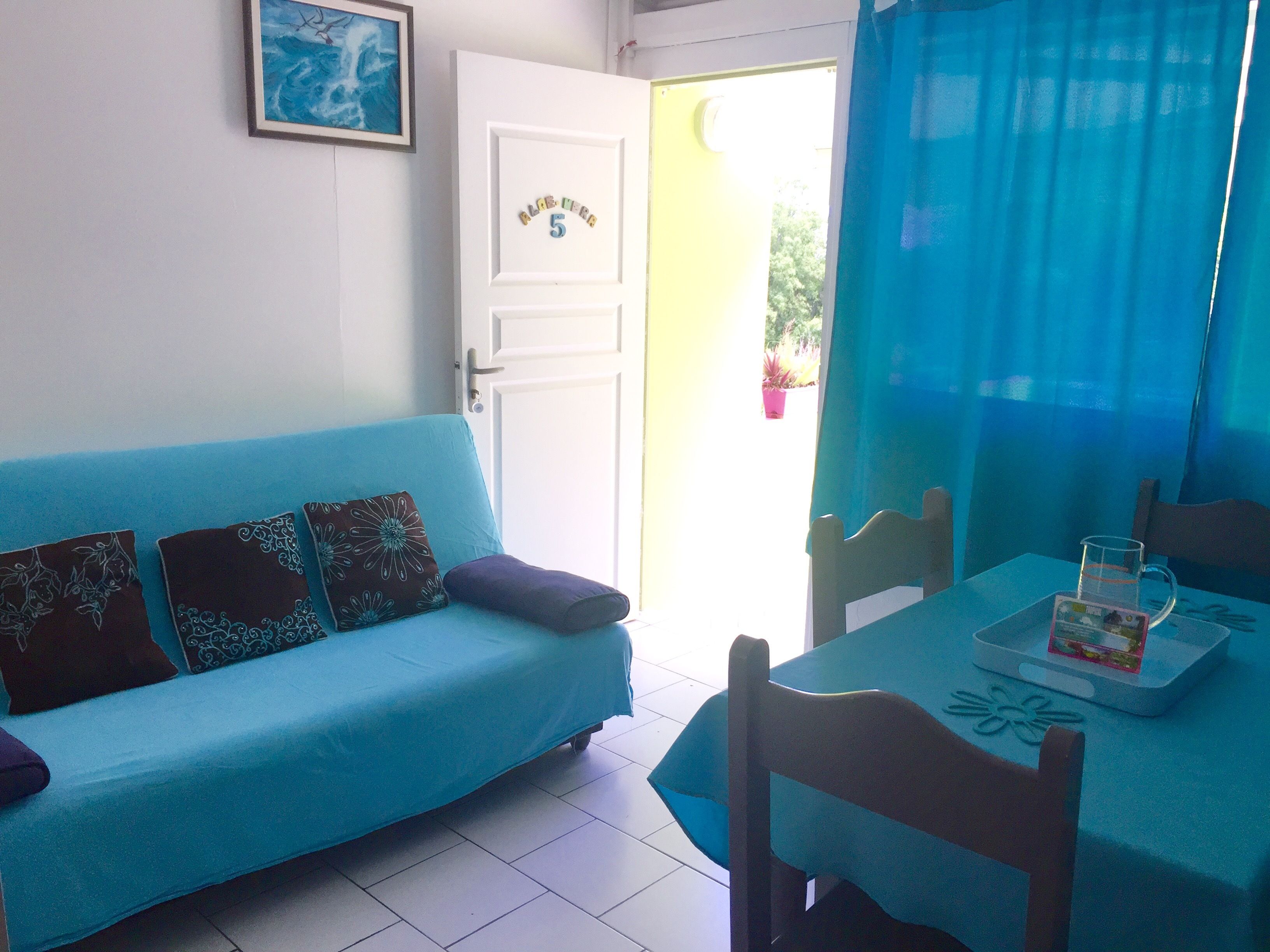Una habitación con un sofá azul, una mesa de comedor con una jarra de vidrio y una puerta con una placa de número.