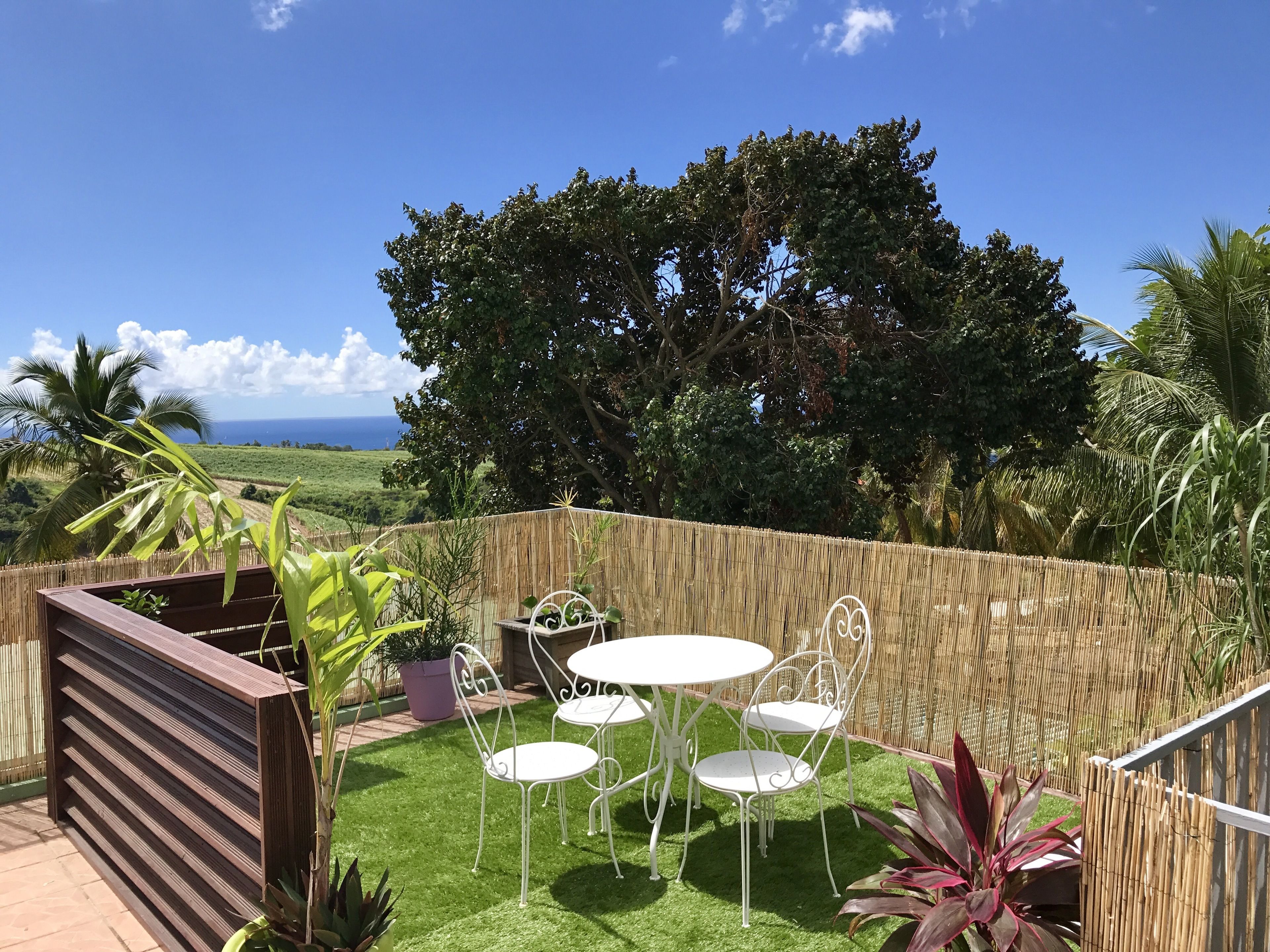 Une terrasse avec une table et des chaises, entourée d’une clôture en bambou, offrant une vue sur l’océan et la végétation.