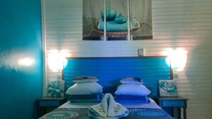 A bedroom with a blue headboard, two bedside tables, a bed with a patterned bedspread, and a painting above the bed. - PARADIS TROPICAL appart'hotel (Baillif)