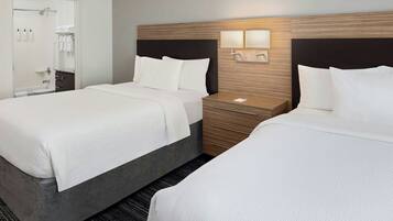Habitación de hotel con dos camas, cabecera de madera, mesita de noche con lámpara y baño visible a través de una puerta abierta.