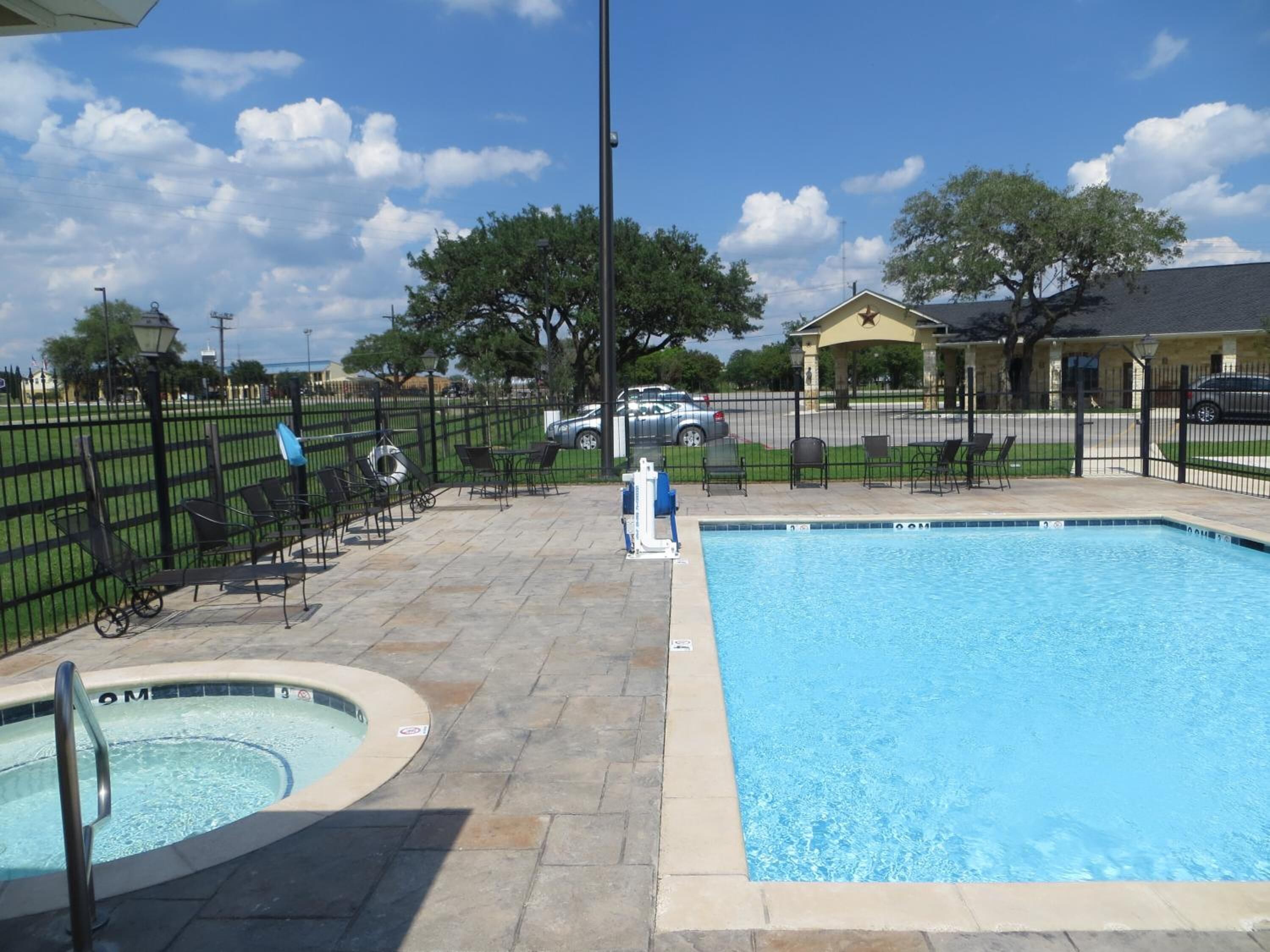 A swimming pool with a hot tub, outdoor seating, and a fenced area with a building in the background.