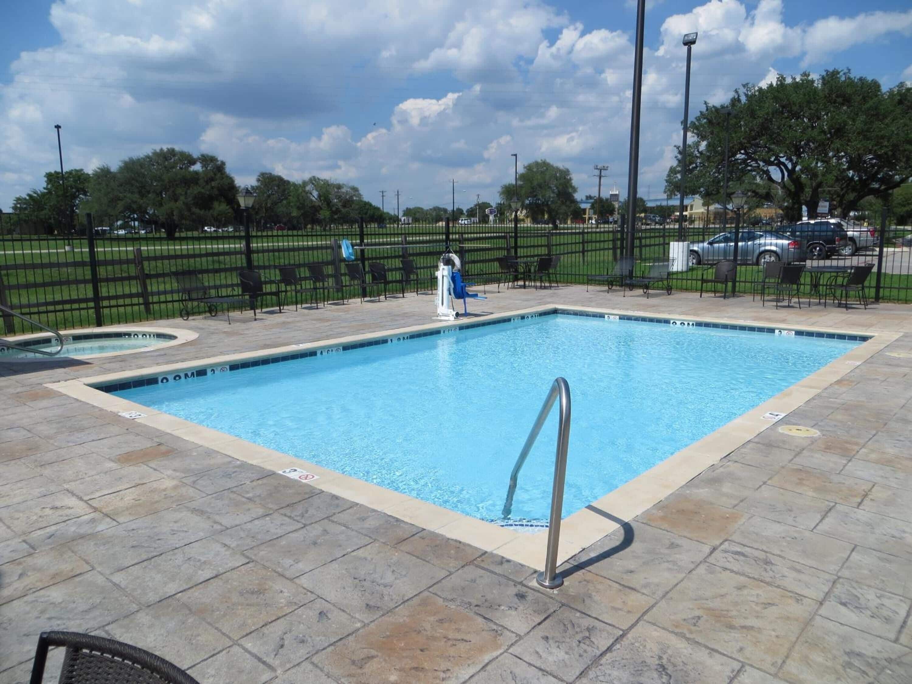 Una piscina rectangular con una escalera metálica, rodeada por un solárium con baldosas y una valla negra.