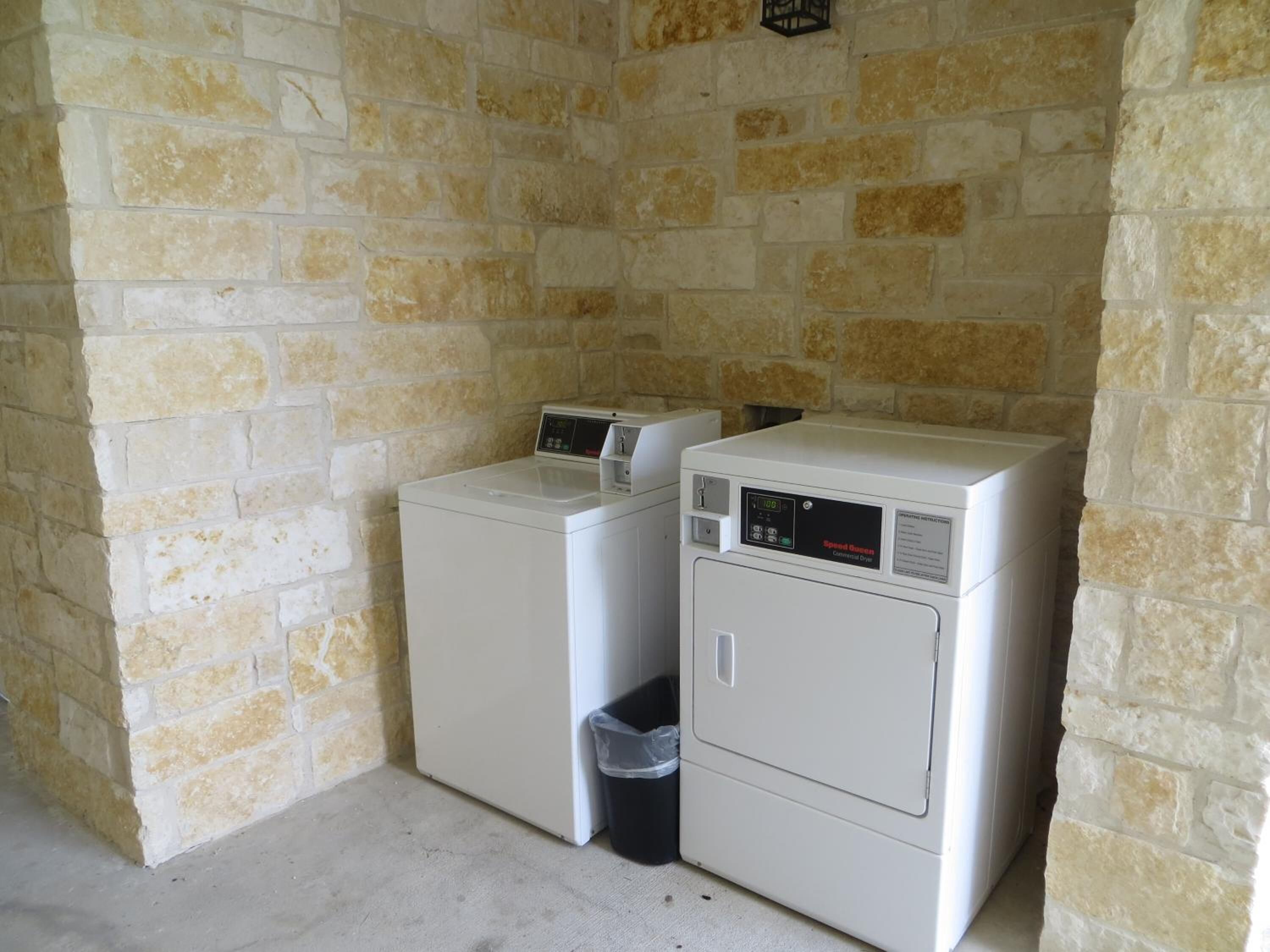 A laundry room with two stacked washers and dryers, a trash bin, and a stone wall.