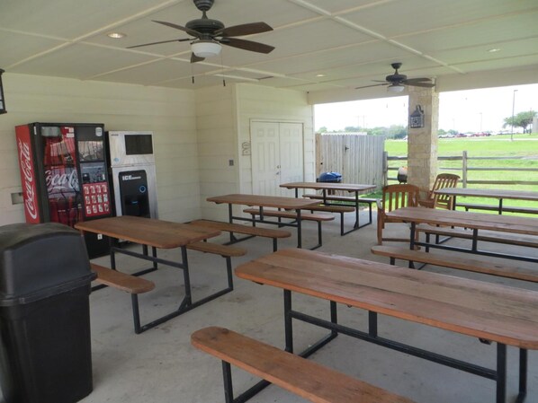 A covered picnic area with tables and benches, a Coca-Cola vending machine, an ice machine, and a view of a grassy field.