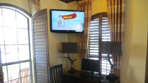 Una habitación con escritorio, dos sillas, un televisor mostrando un anuncio y una ventana con persianas.