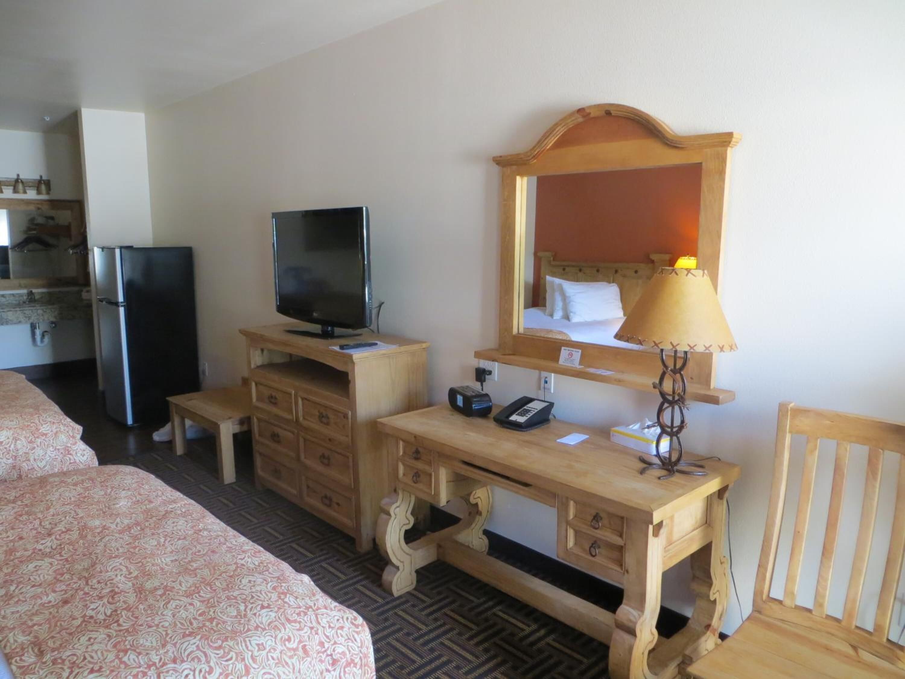Una habitación de hotel con cama, escritorio de madera, televisor, refrigerador, espejo y lámpara.