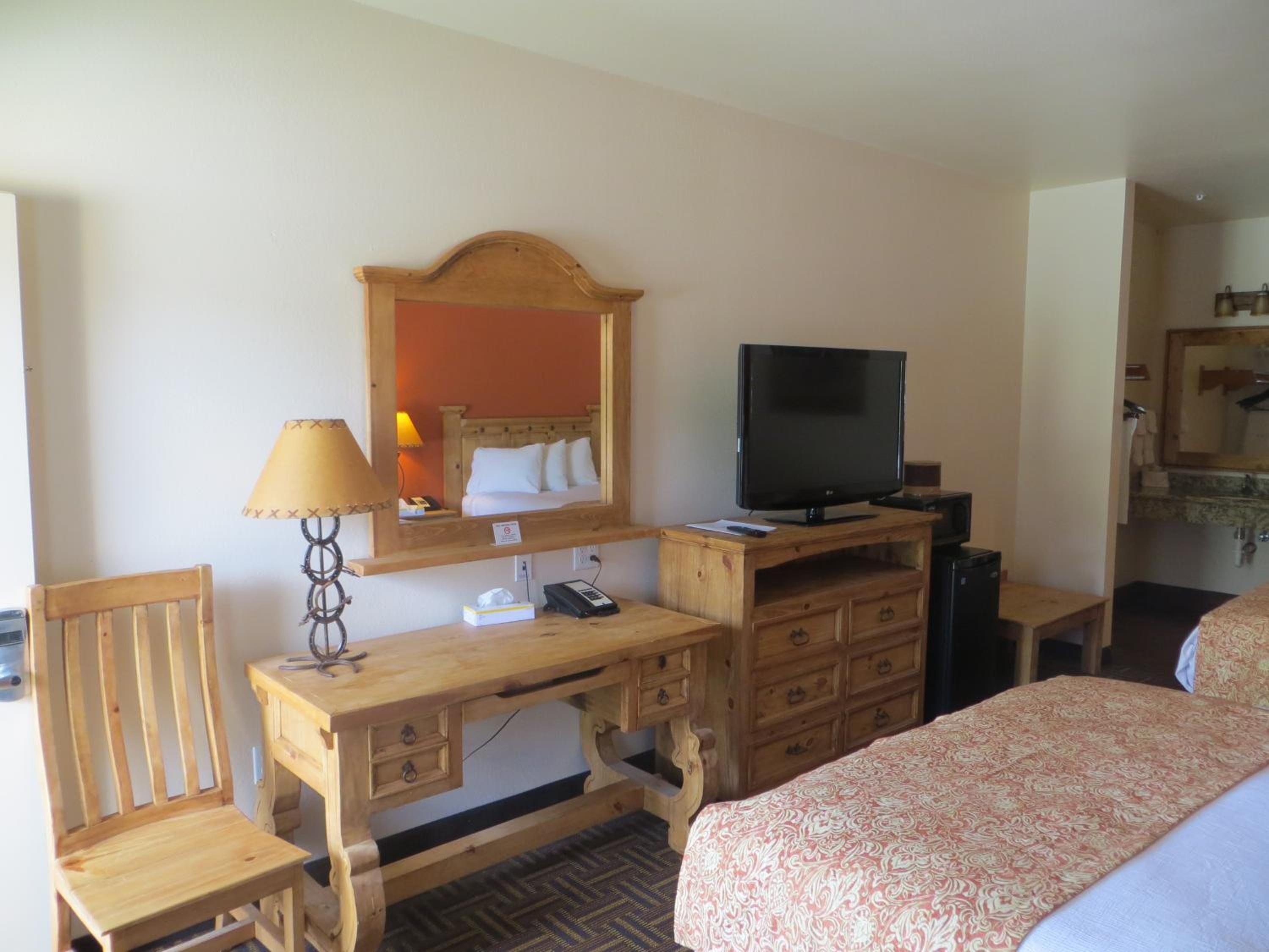 A hotel room with a wooden desk, a television, a bed with a patterned bedspread, a chair, and a mirror.