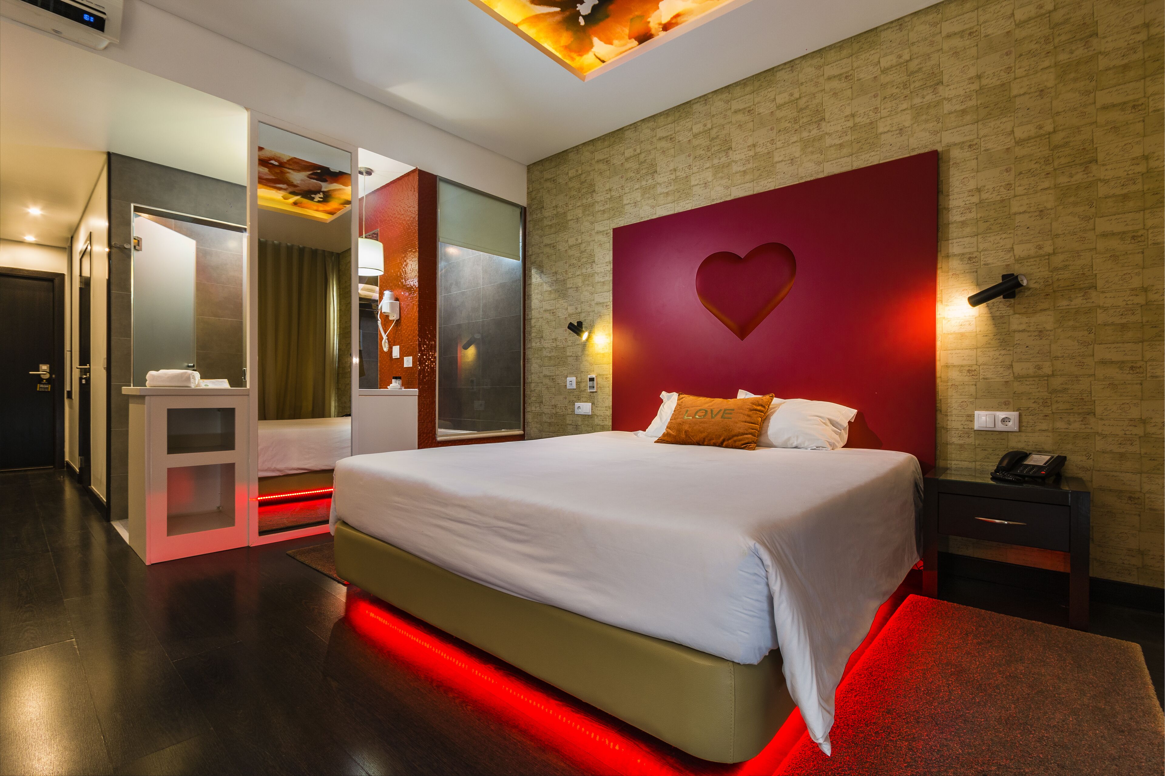 A modern hotel room with a large bed, a red accent wall with a heart design, a small bedside table with a phone, and a separate bathroom area.