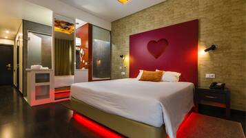 A modern hotel room with a large bed, a red accent wall with a heart design, a small bedside table with a phone, and a separate bathroom area.