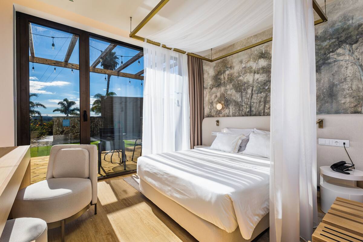 Un dormitorio moderno con una cama grande, un escritorio y vistas a palmeras a través de amplias ventanas.