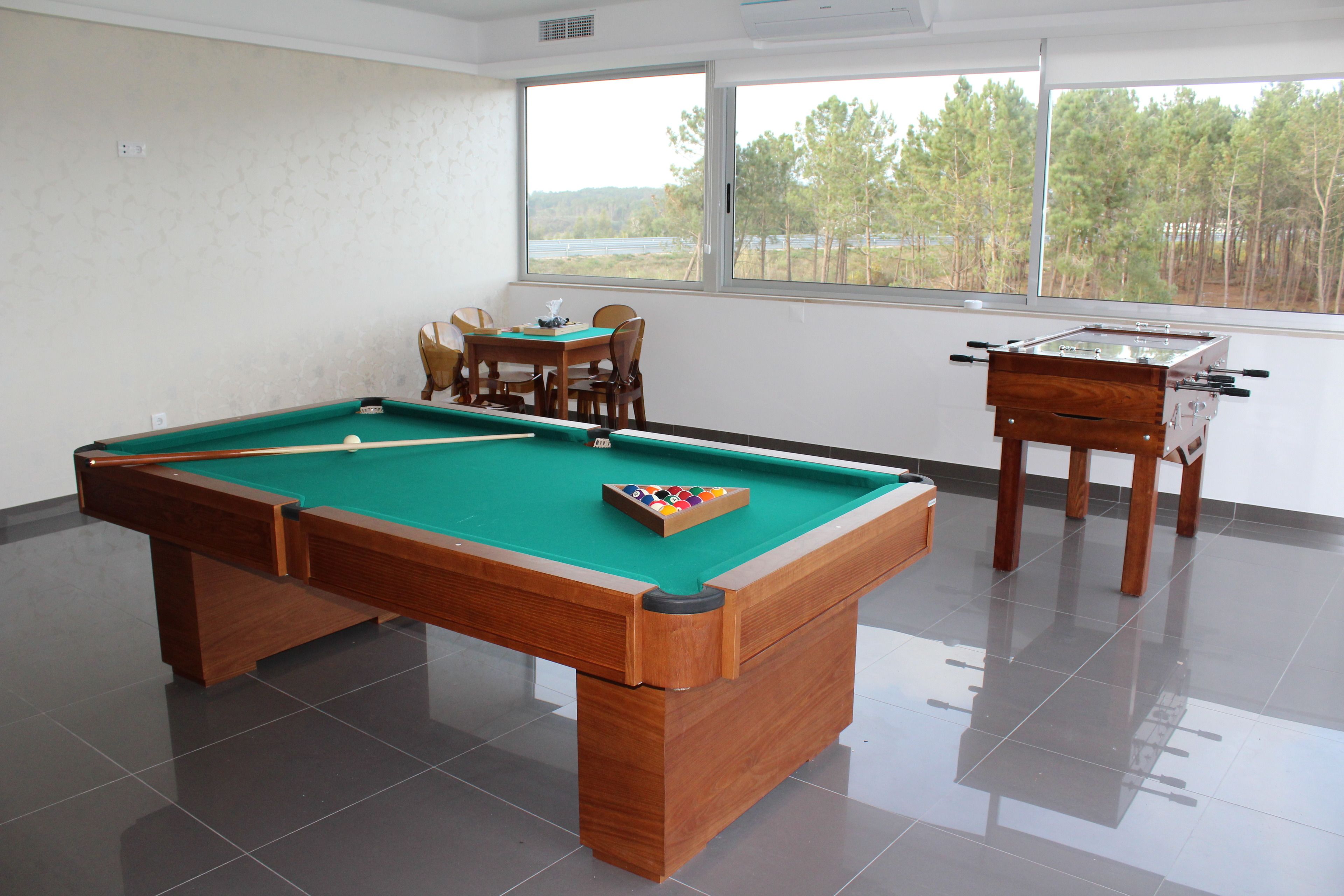 Una mesa de billar junto a una mesa de fútbol de mesa y sillas en una habitación con ventanales amplios.