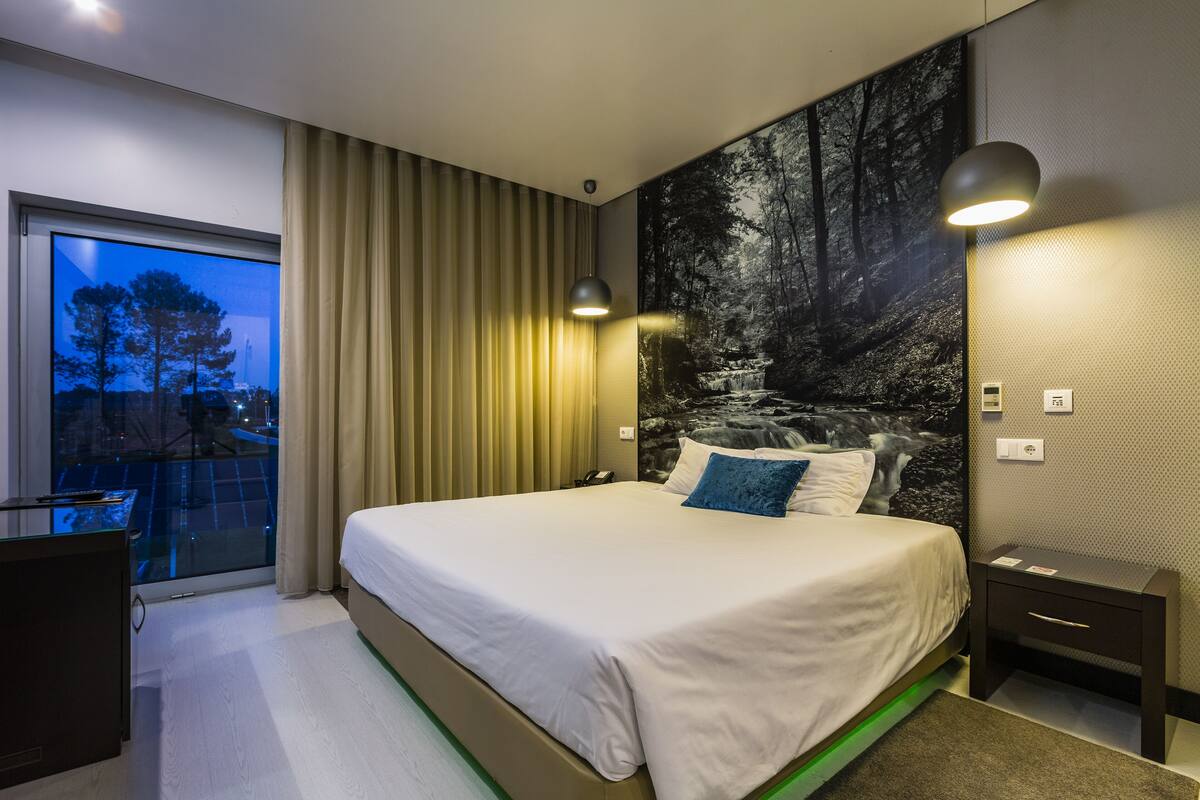 Habitación de hotel moderna con una cama grande, un escritorio y vistas a un paisaje urbano a través de un ventanal.
