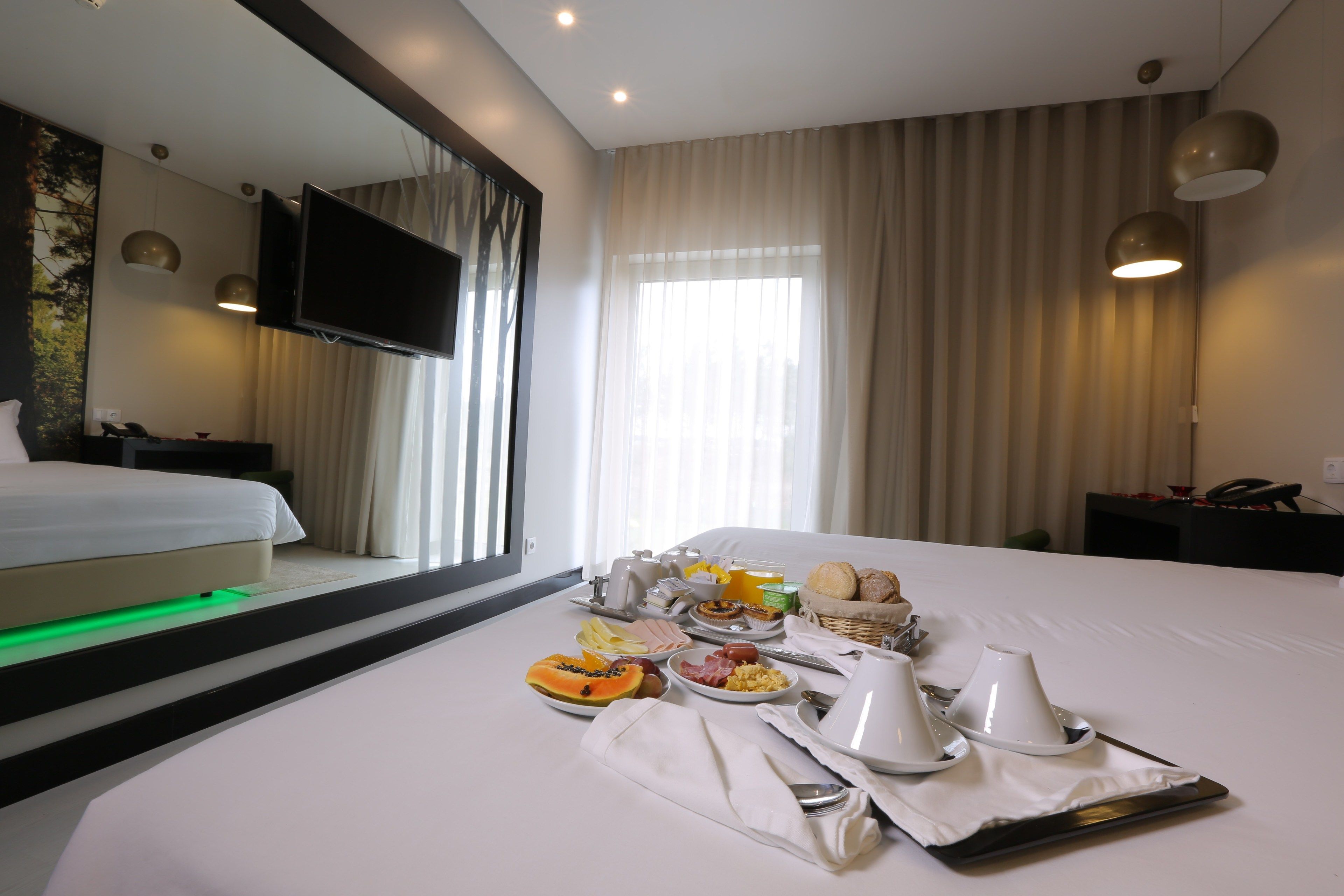 A hotel room with a neatly made bed, breakfast tray with fruits and eggs, a flat-screen TV, and a large window with curtains.