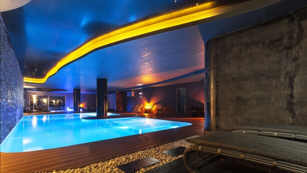 A modern indoor pool area with a wooden deck, stone pathway, and lounge chairs.