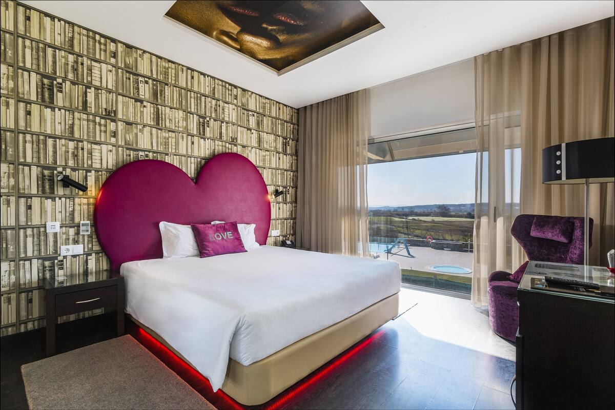 Una habitación de hotel moderna con una cama grande, un cabecero con forma de corazón, vistas al exterior y un escritorio de vidrio.
