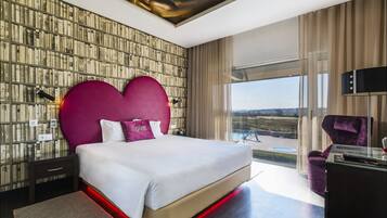 Una habitación de hotel moderna con una cama grande, un cabecero con forma de corazón, vistas al exterior y un escritorio de vidrio.