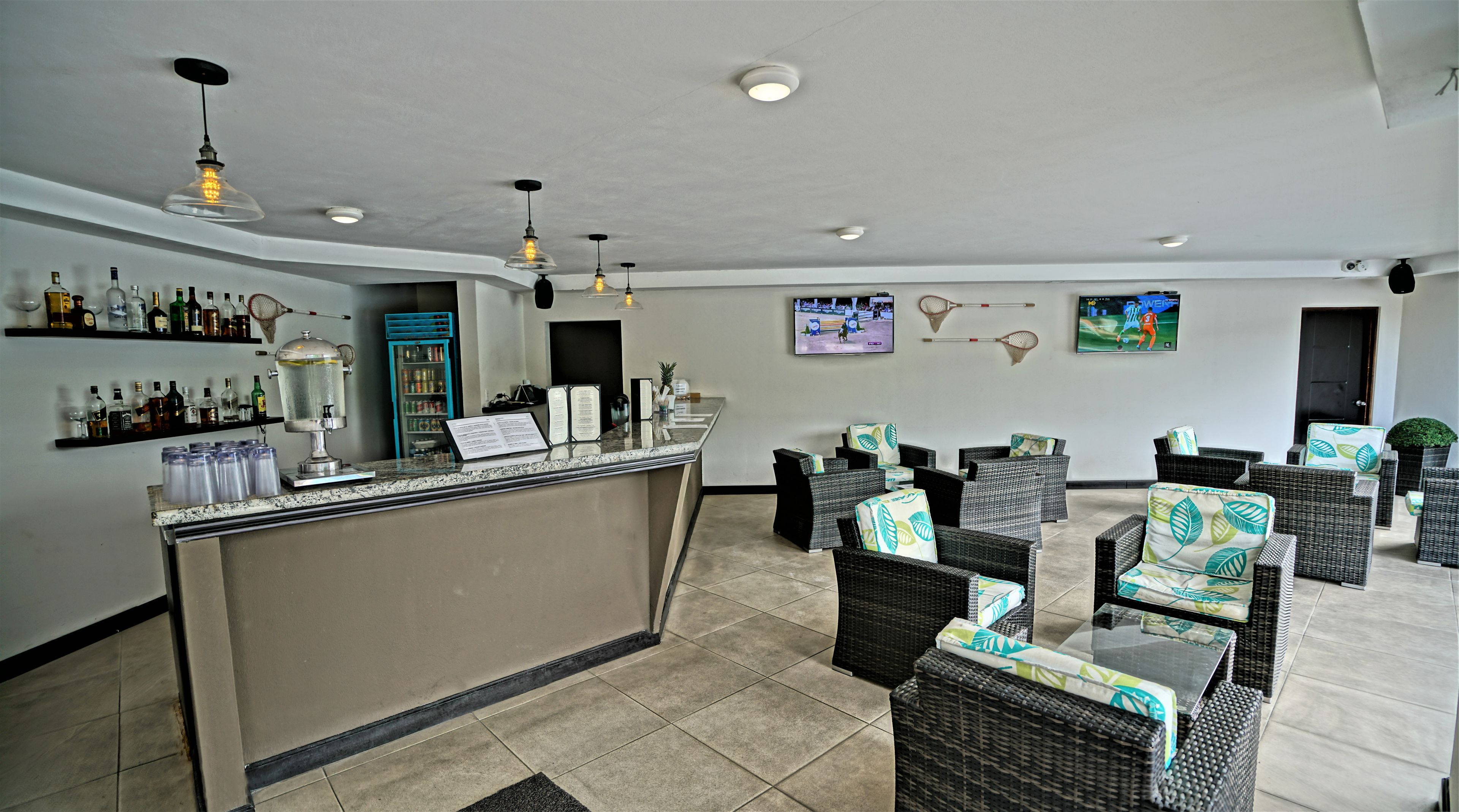 A hotel lobby with a reception desk, wicker furniture, a beverage bar, and a TV screen displaying a sports event.