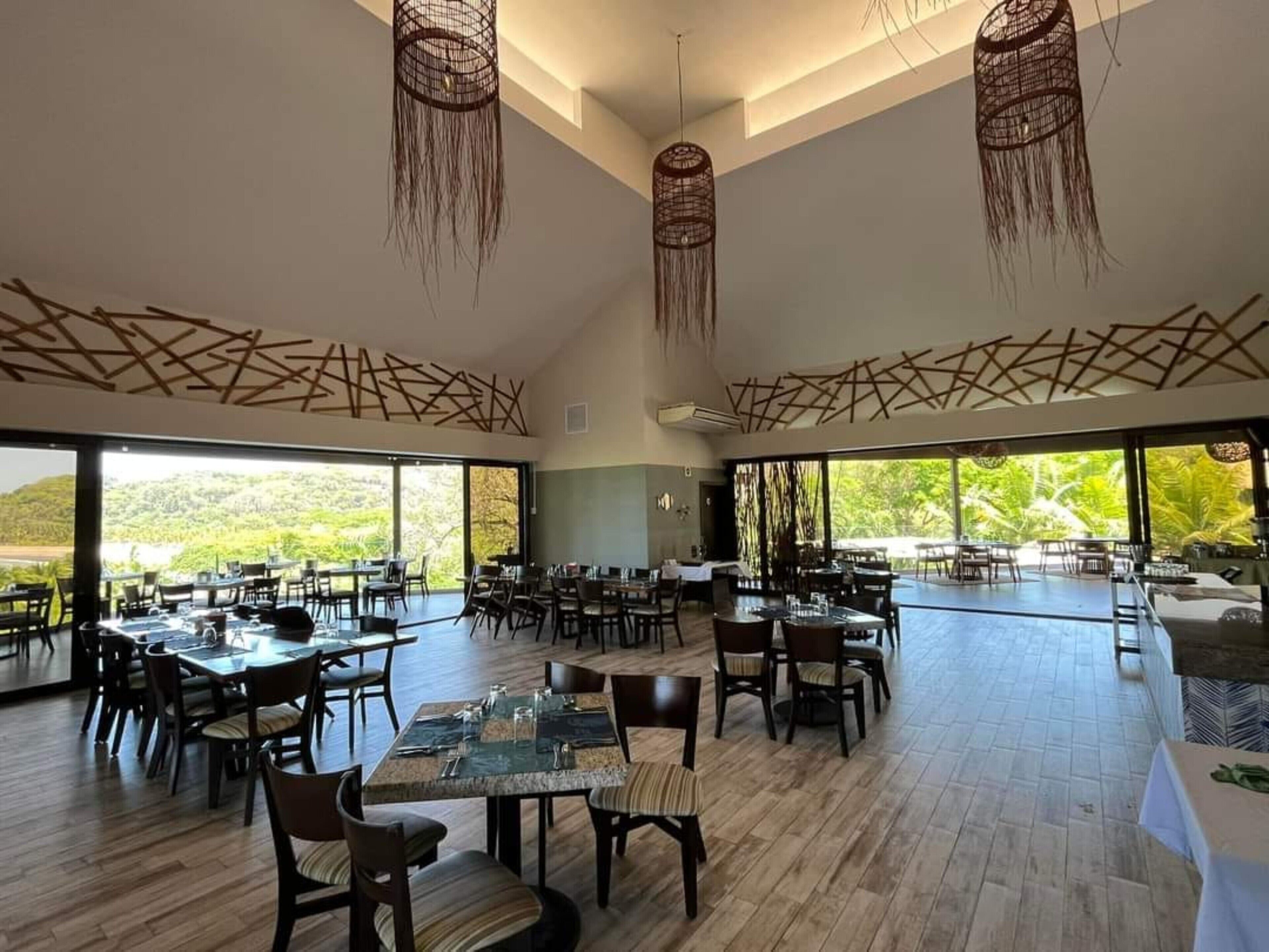 Un comedor espacioso con techos altos, ventanas grandes y lámparas colgantes. Las mesas están dispuestas para comer, y la habitación presenta un diseño moderno y limpio.