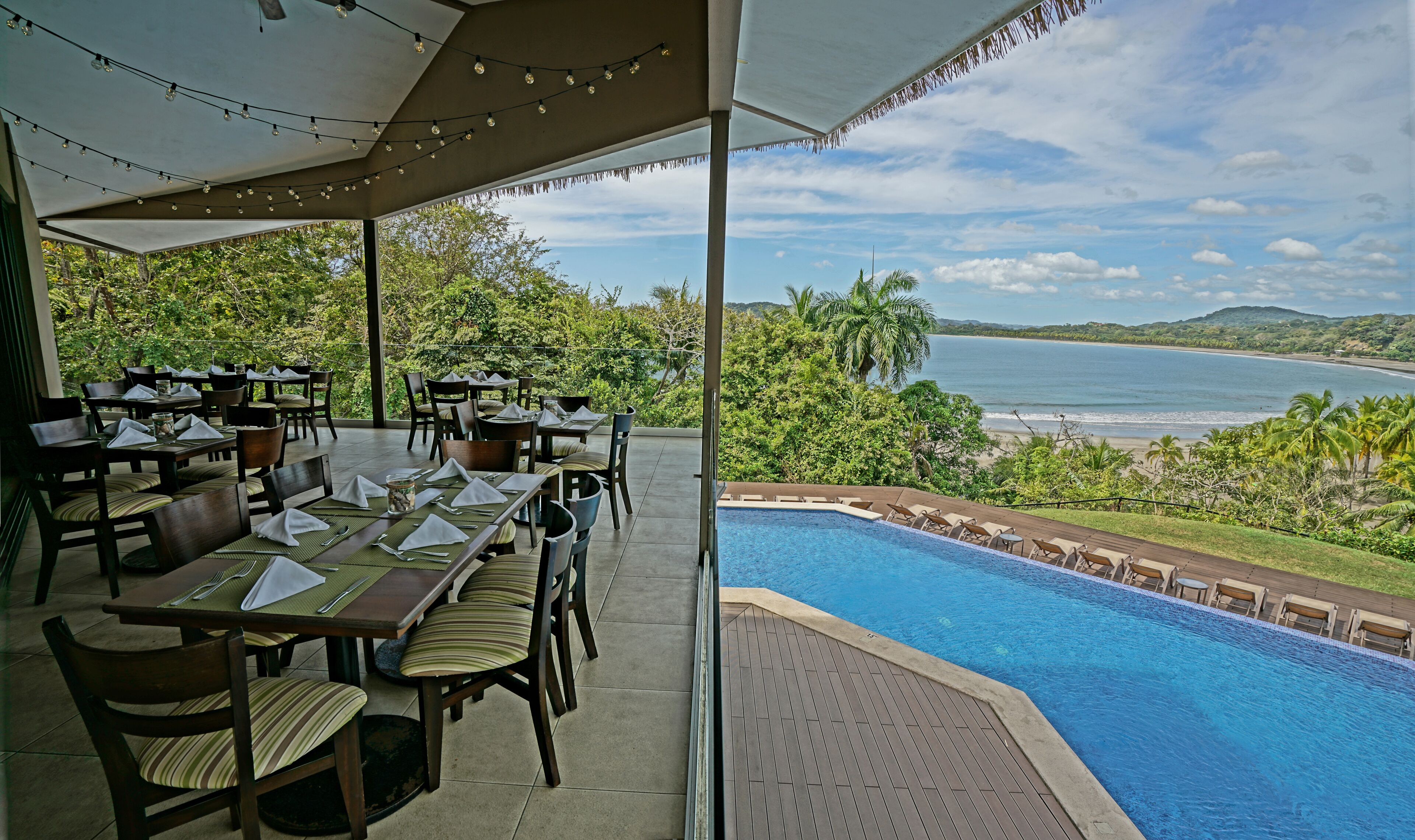Un espacio de comedor al aire libre con mesas y sillas, con vistas a una piscina y una playa.