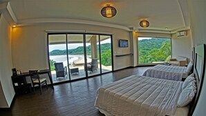 Un dormitorio con dos camas, un escritorio y una silla, con un amplio ventanal corredero que da a un balcón con vistas a la playa y las montañas.