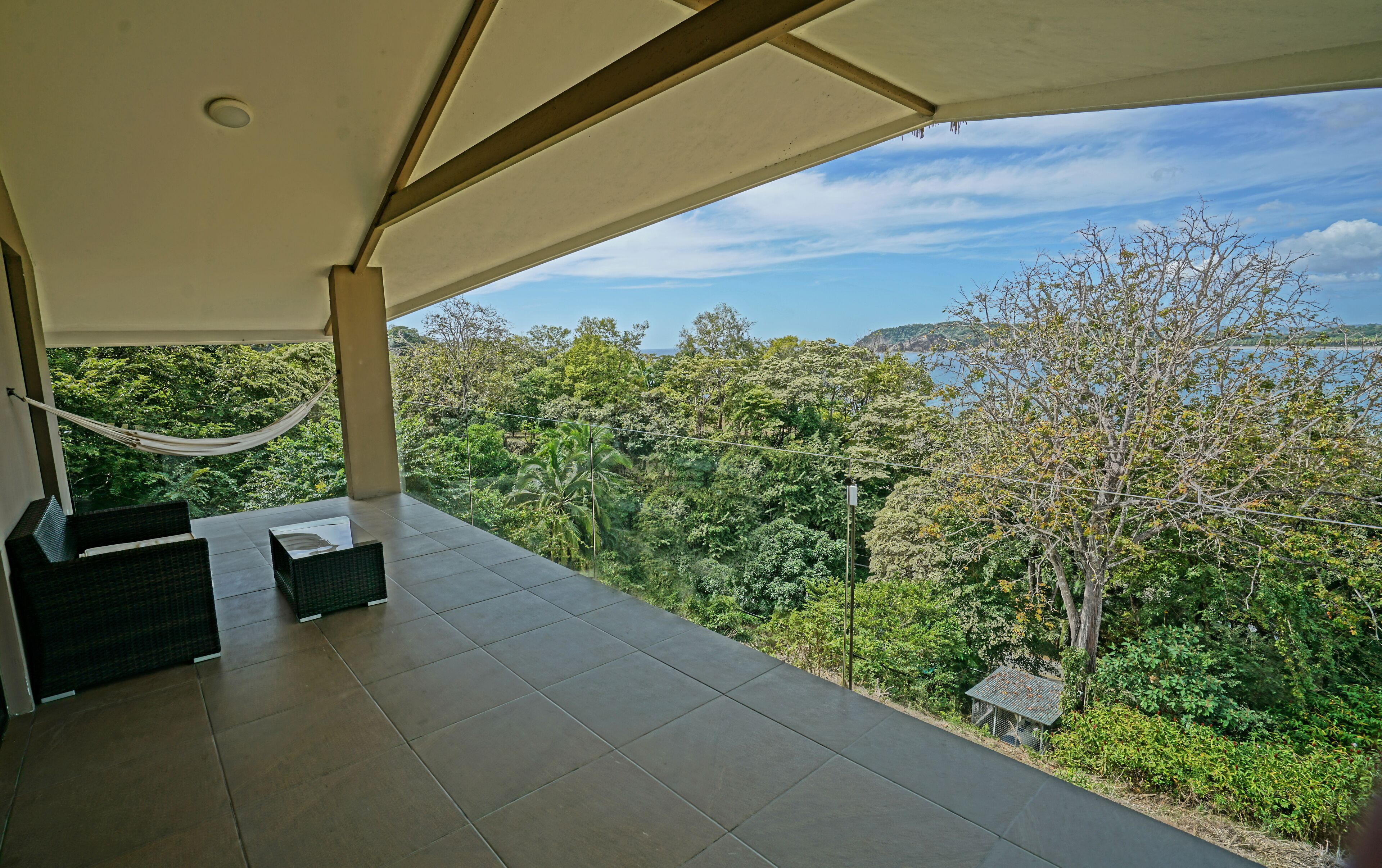 A covered balcony with a hammock, two wicker chairs, and a view of a lush forest and coastline.