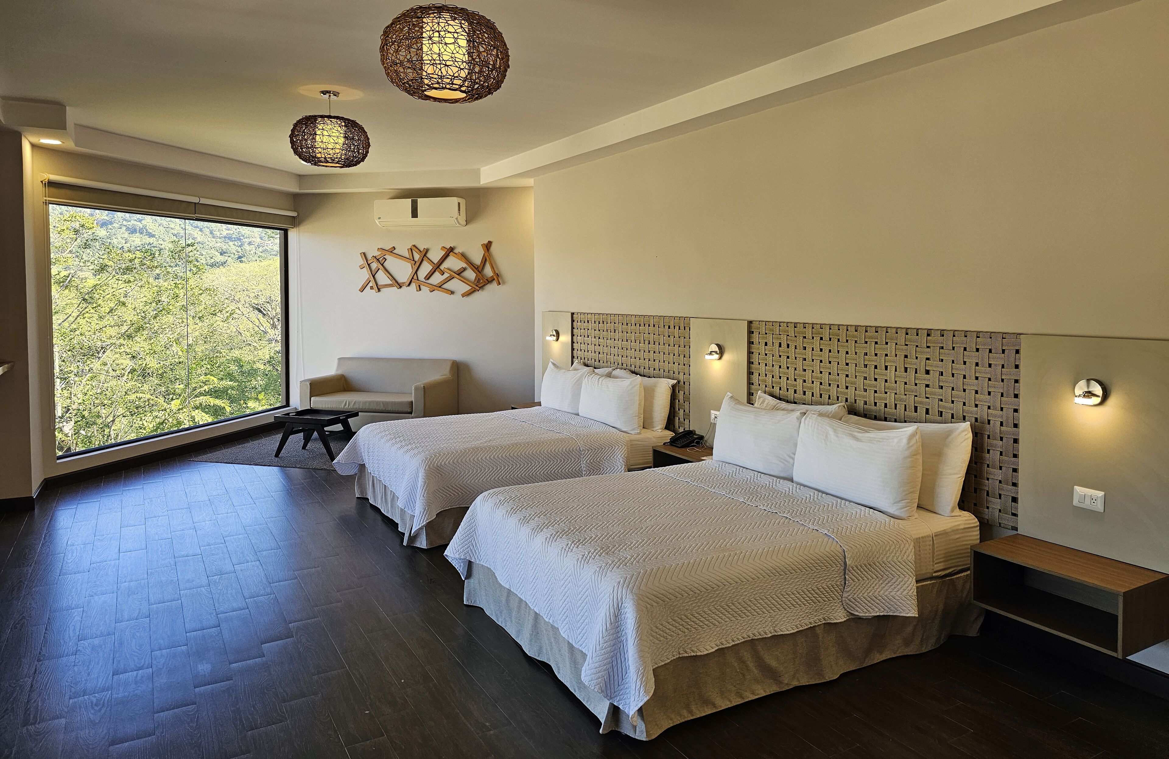 A modern hotel room with two beds, a large window with a view of trees, and a woven light fixture.
