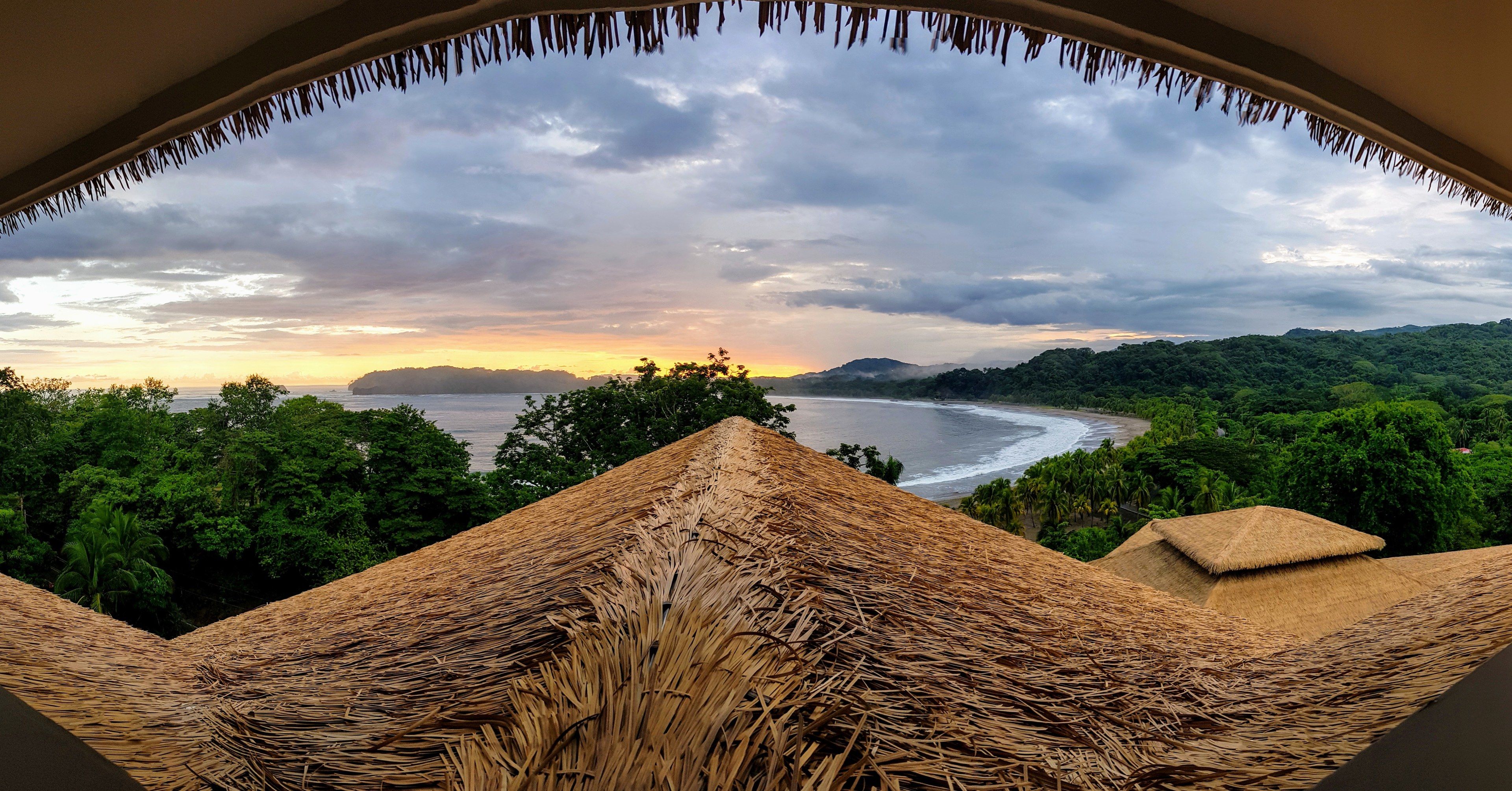 Vista desde una estructura con techo de paja que domina una playa con abundante vegetación y un cielo nublado.