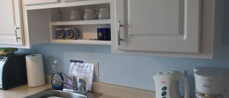 A kitchen with white cabinets, a sink with a dish rack, a coffee maker, and a kettle.