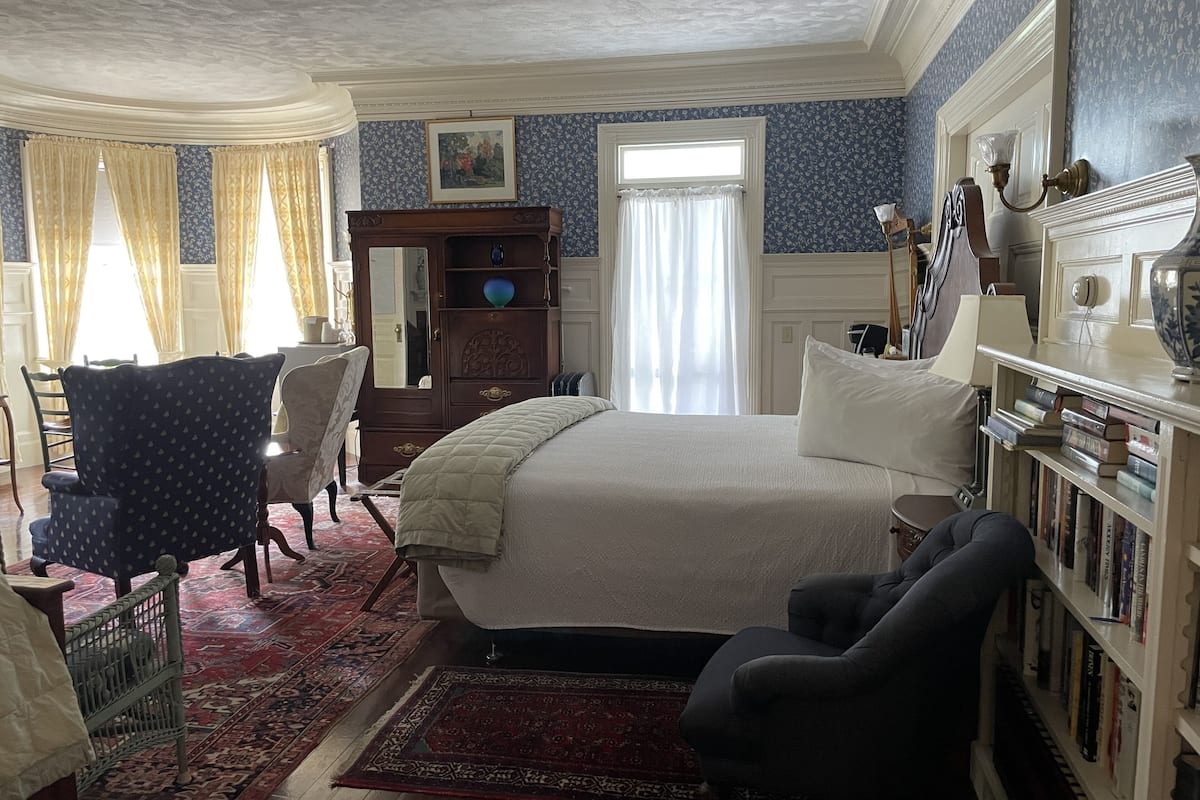 A bedroom with a large bed, a wooden dresser, a chair, a bookshelf, and a window with curtains.