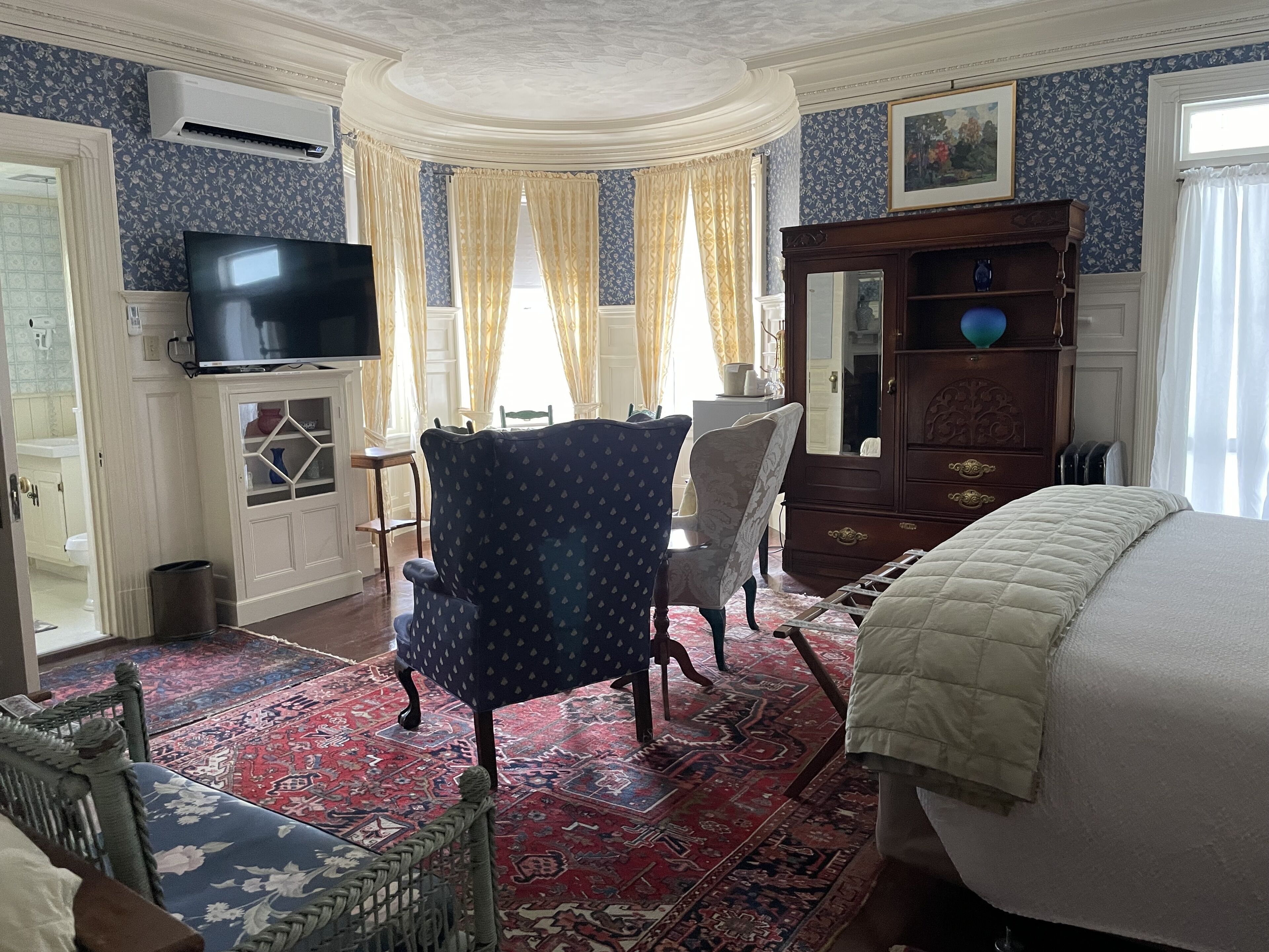 A room with a television, a wooden cabinet, a chair, a bed, and a patterned rug.