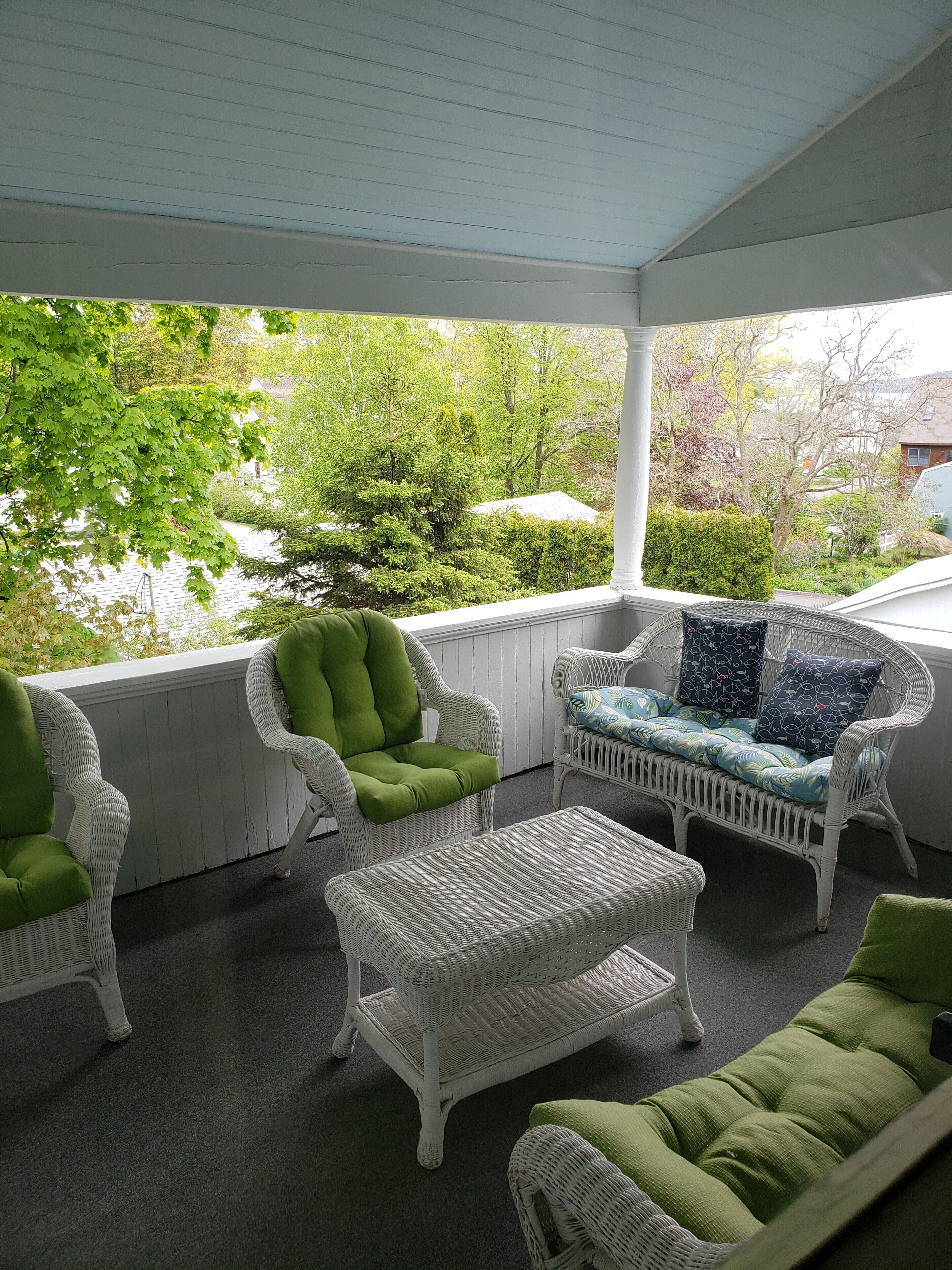 A covered porch with wicker furniture, including chairs and a sofa, and a view of greenery and trees.