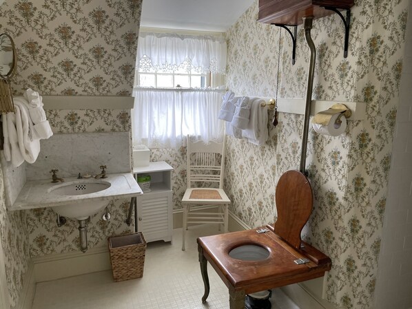 Baño con inodoro de madera, lavamanos y espejo. Se ve una ventana con cortinas, una silla y toallas colgadas en un perchero.