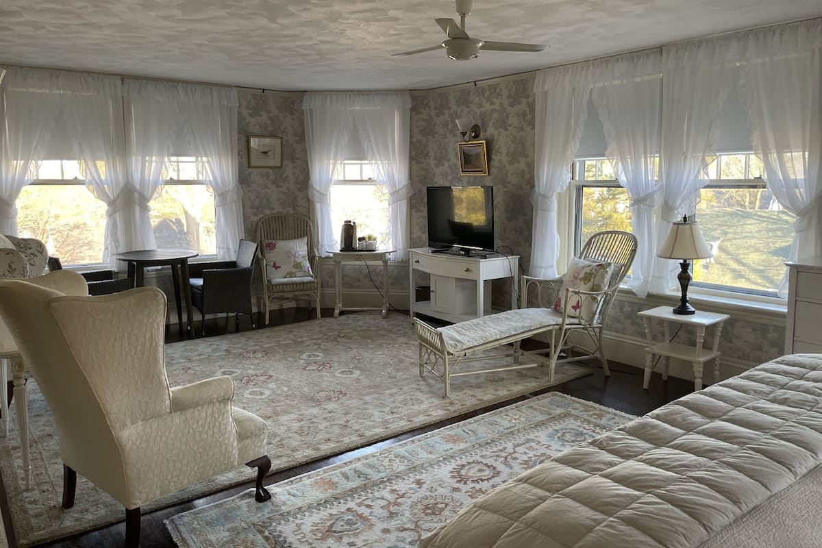 A room with a bed, two chairs, a desk with a TV, a lamp, and a window with curtains.