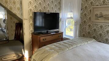 Une chambre à coucher avec un lit, un téléviseur posé sur une commode en bois, une fenêtre avec des rideaux et une porte donnant sur une autre pièce.