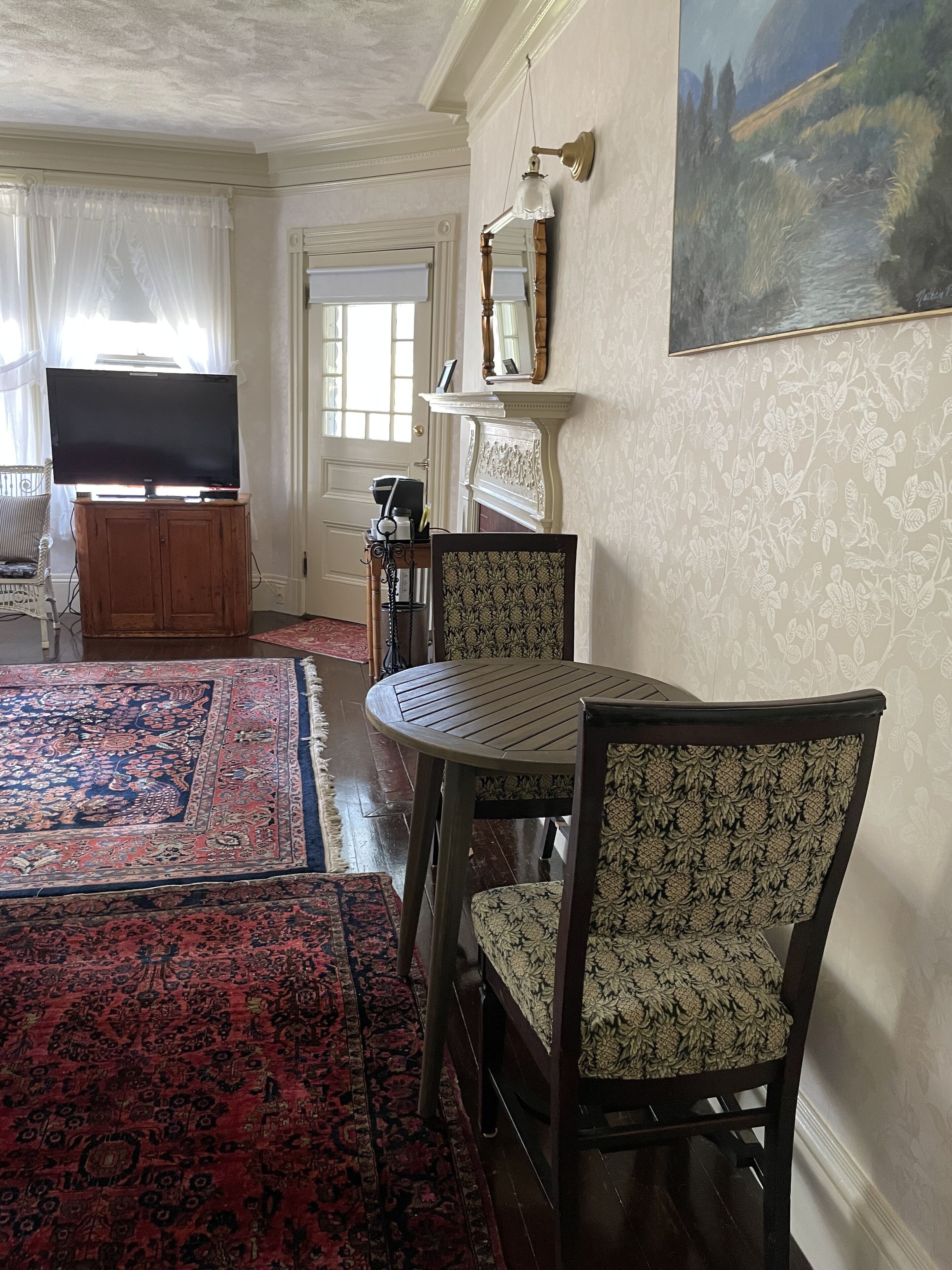 Un salon avec une table ronde, deux fauteuils à motifs, un téléviseur posé sur un meuble, une cheminée avec un miroir et un tableau accroché au mur.