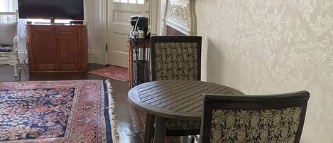 Un salon avec une table ronde, deux fauteuils à motifs, un téléviseur posé sur un meuble, une cheminée avec un miroir et un tableau accroché au mur.