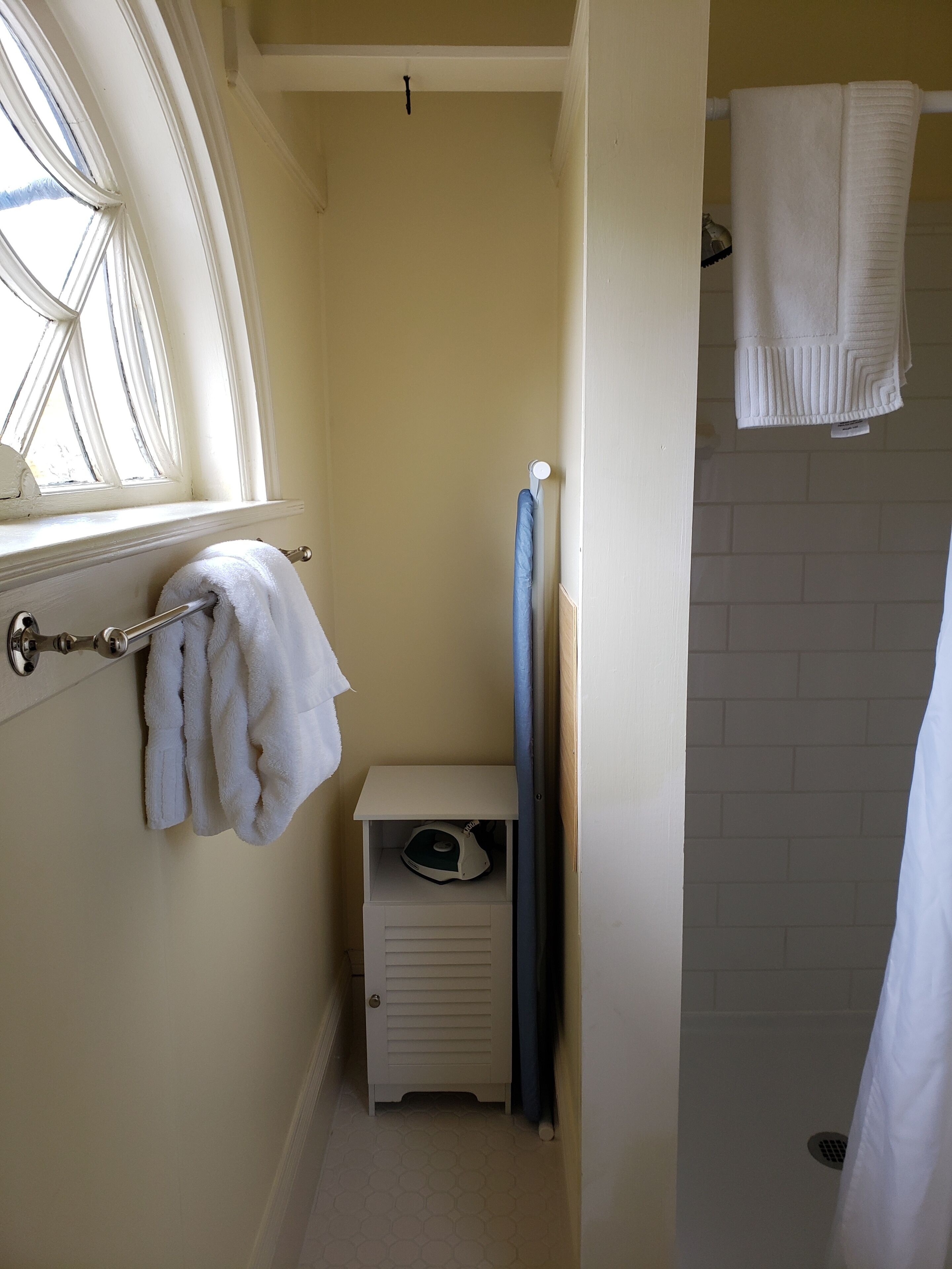 A bathroom with a window, a towel rack, a small shelf with a hairdryer, and a shower area.