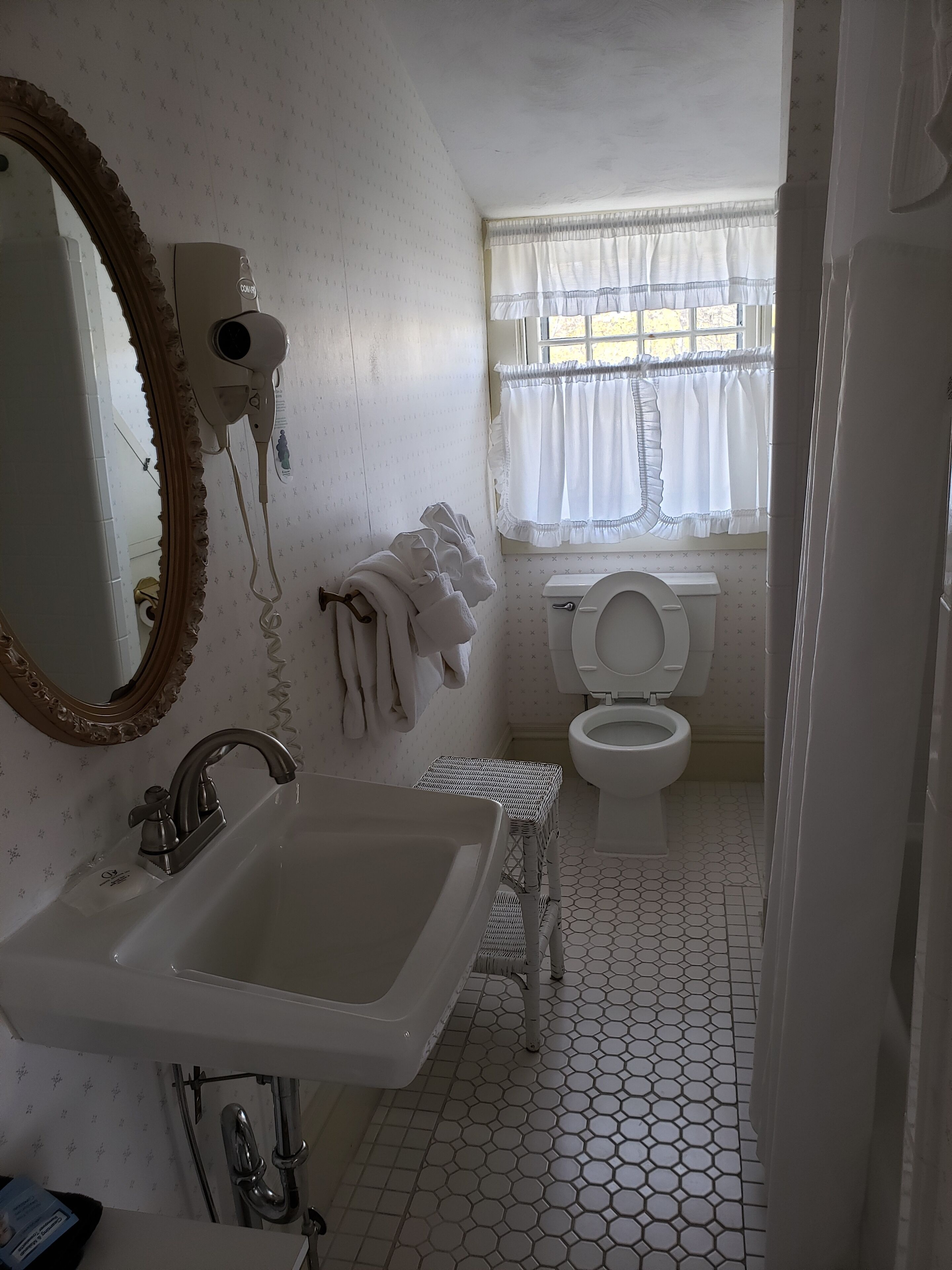 A bathroom with a sink, mirror, hairdryer, toilet, and window with curtains.