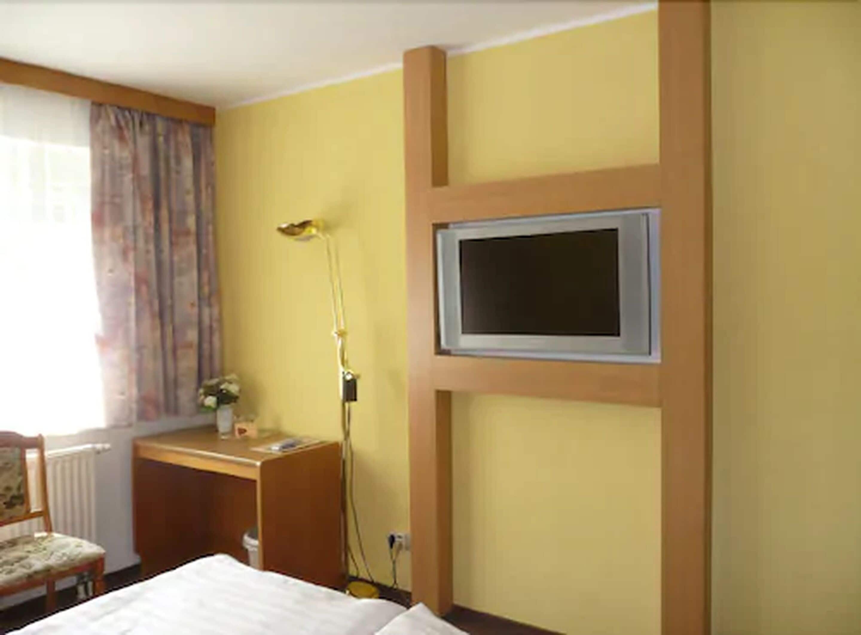 A hotel room with a bed, a desk, a chair, a television mounted on the wall, and a window with curtains.