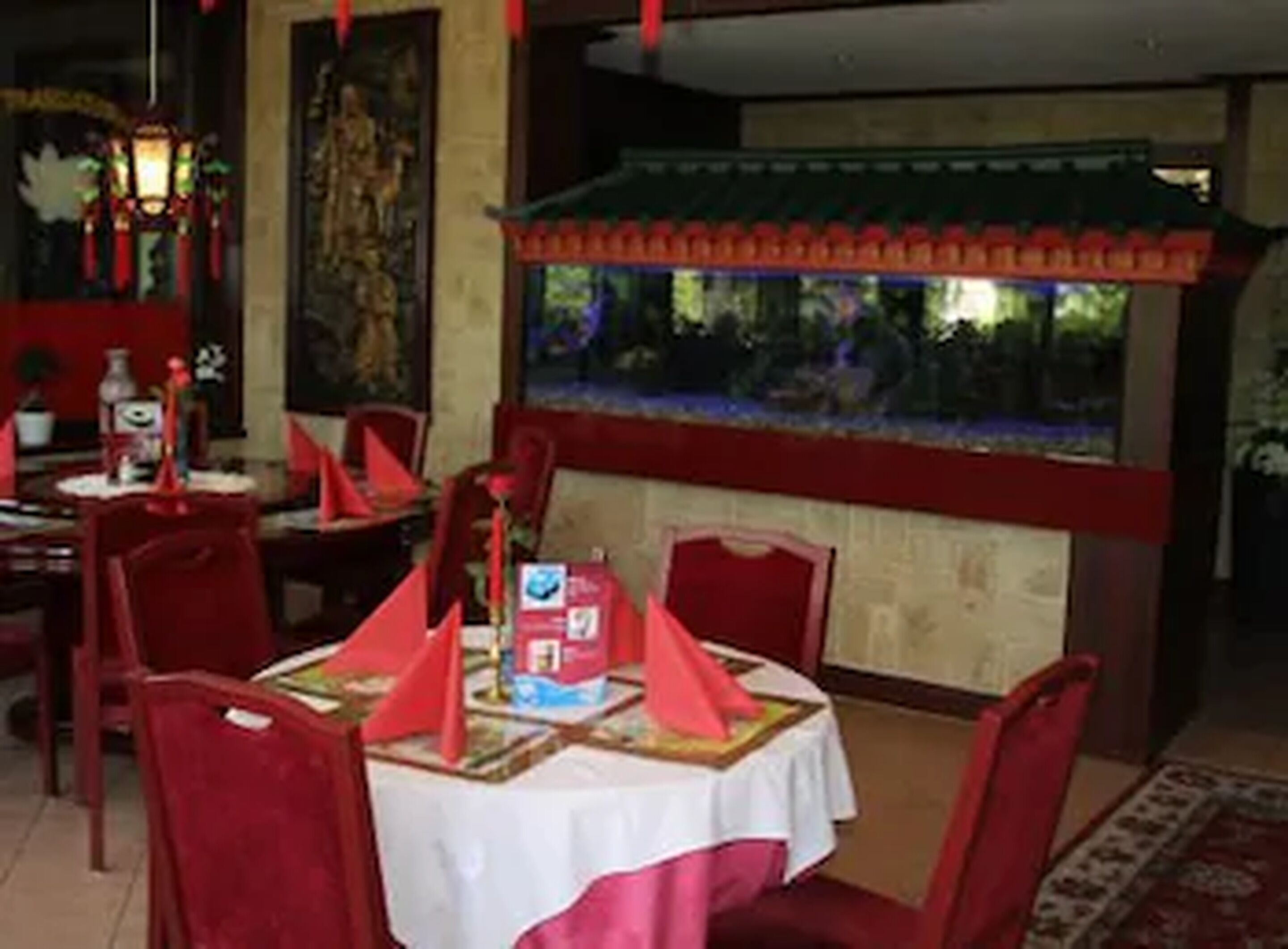 A dining area with red chairs and a table set for two, featuring a traditional Chinese decorative lantern and a large fish tank in the background.