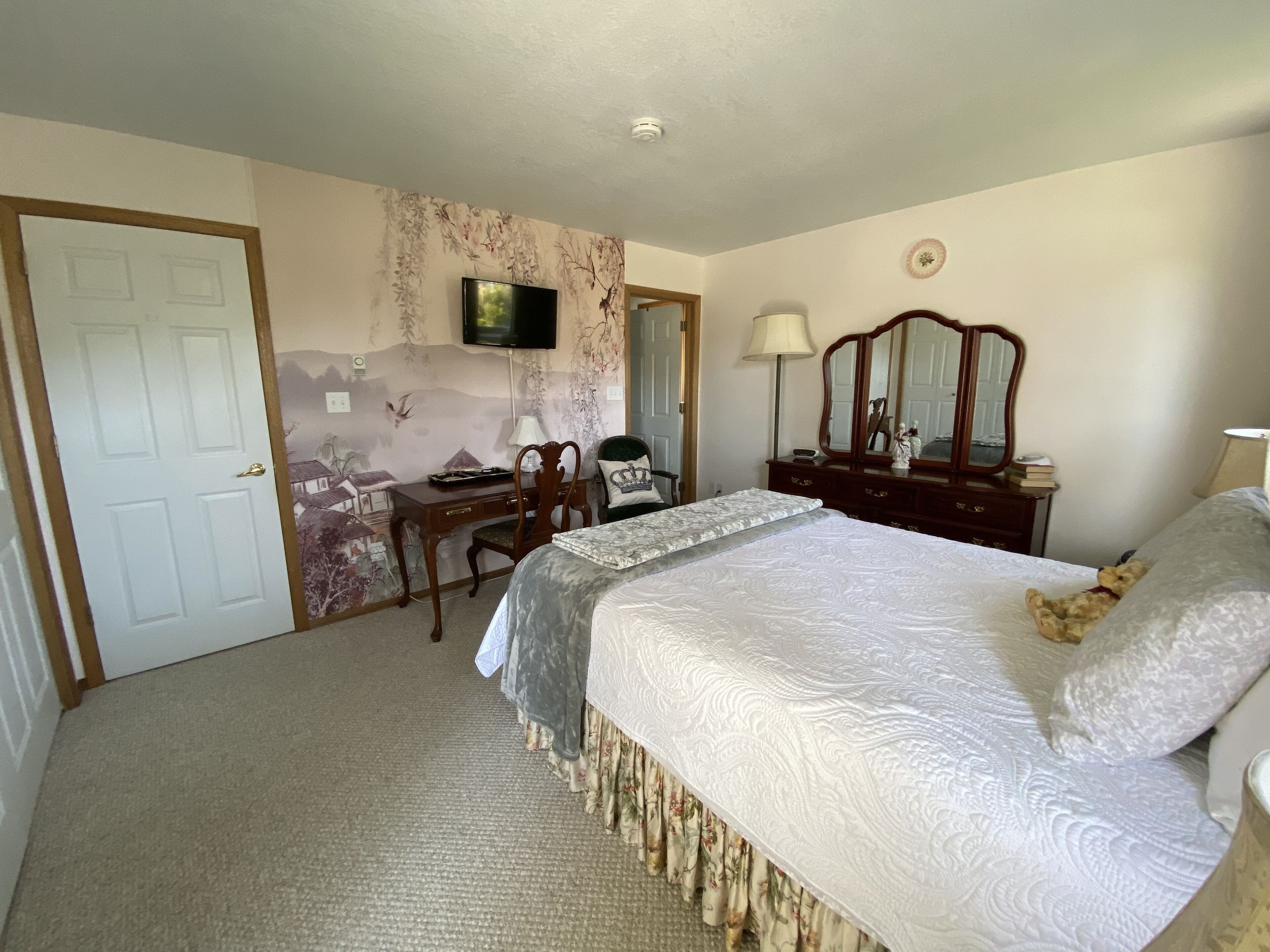 Une chambre à coucher comprenant un lit, un bureau, une chaise, une commode, un miroir et un téléviseur fixé au mur.