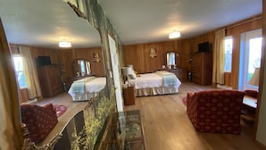 Una habitación con paredes y suelos de madera, dos camas con ropa de cama blanca, un sillón rojo con estampado, un armario de madera y un espejo grande.
