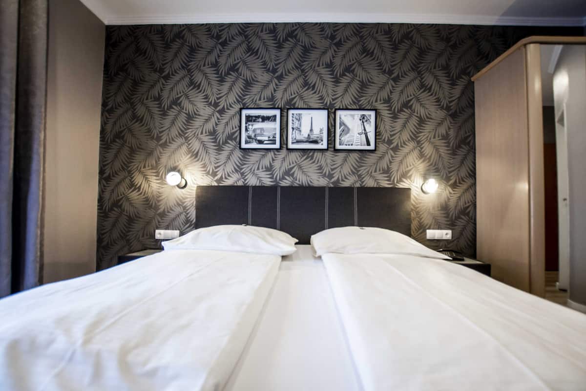Uma cama de casal com roupa de cama branca, papel de parede com padrão de folhagens e três quadros na parede.