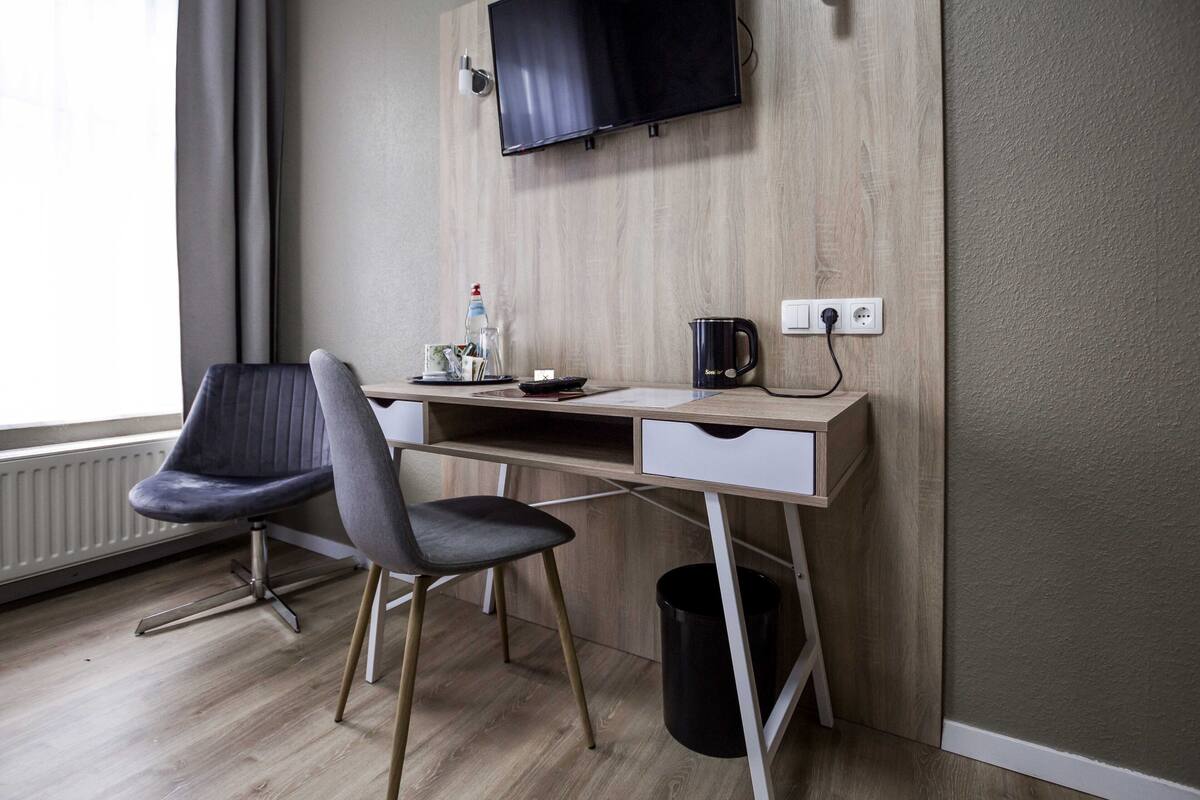 Quarto moderno com escrivaninha de madeira, uma cadeira cinza, televisão fixada na parede e radiador.